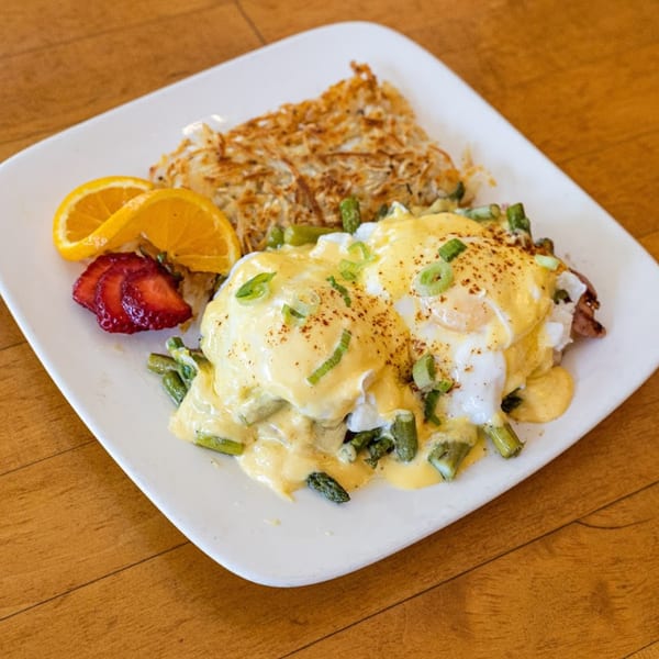 Eggs Benedict with asparagus, hash browns, orange slices, and strawberries on a white plate.