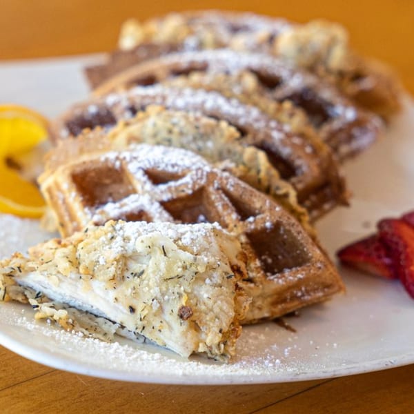 Chicken and waffles with powdered sugar, lemon, and strawberries on a white plate.