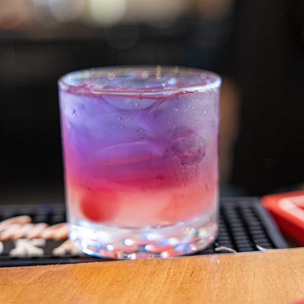 Vibrant purple and pink layered cocktail with ice in a clear glass on a wooden counter.