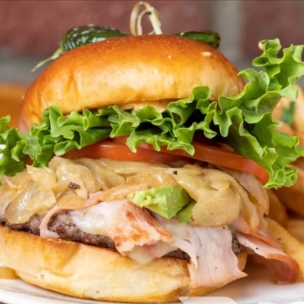 A burger with lettuce, tomato, caramelized onions, bacon, avocado, and a pickle on top.